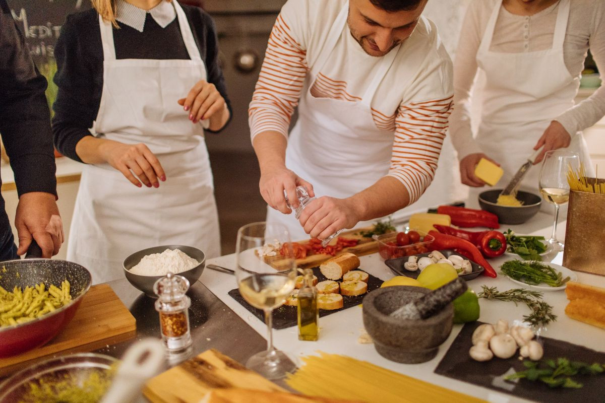 A group of people participating in a cooking class, wearing aprons and working together on a variety of fresh ingredients, including vegetables, pasta, and bread. One person is grating cheese while another is seasoning a dish, creating a lively and collaborative atmosphere in the kitchen.